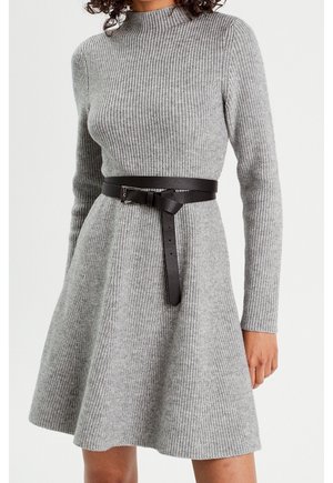 Femme portant une robe gris clair à manches longues en maille côtelée, avec une ceinture en cuir noir nouée à la taille.