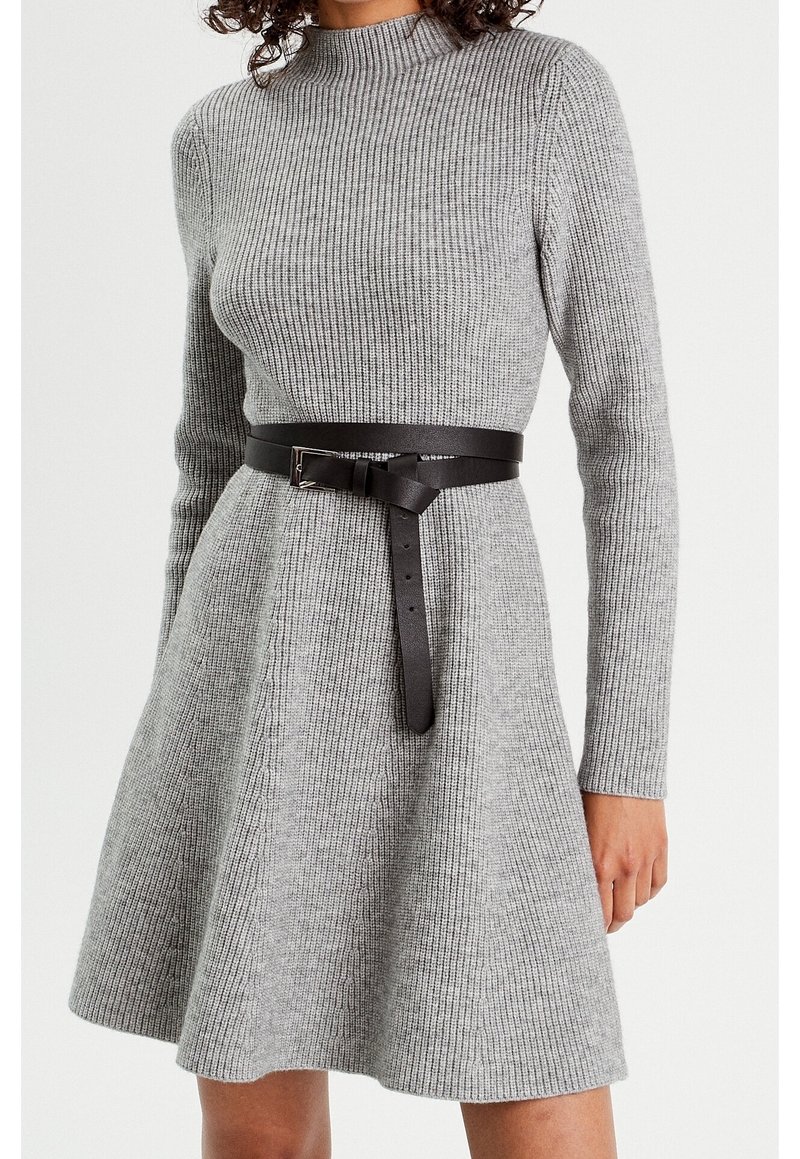 Femme portant une robe gris clair à manches longues en maille côtelée, avec une ceinture en cuir noir nouée à la taille.