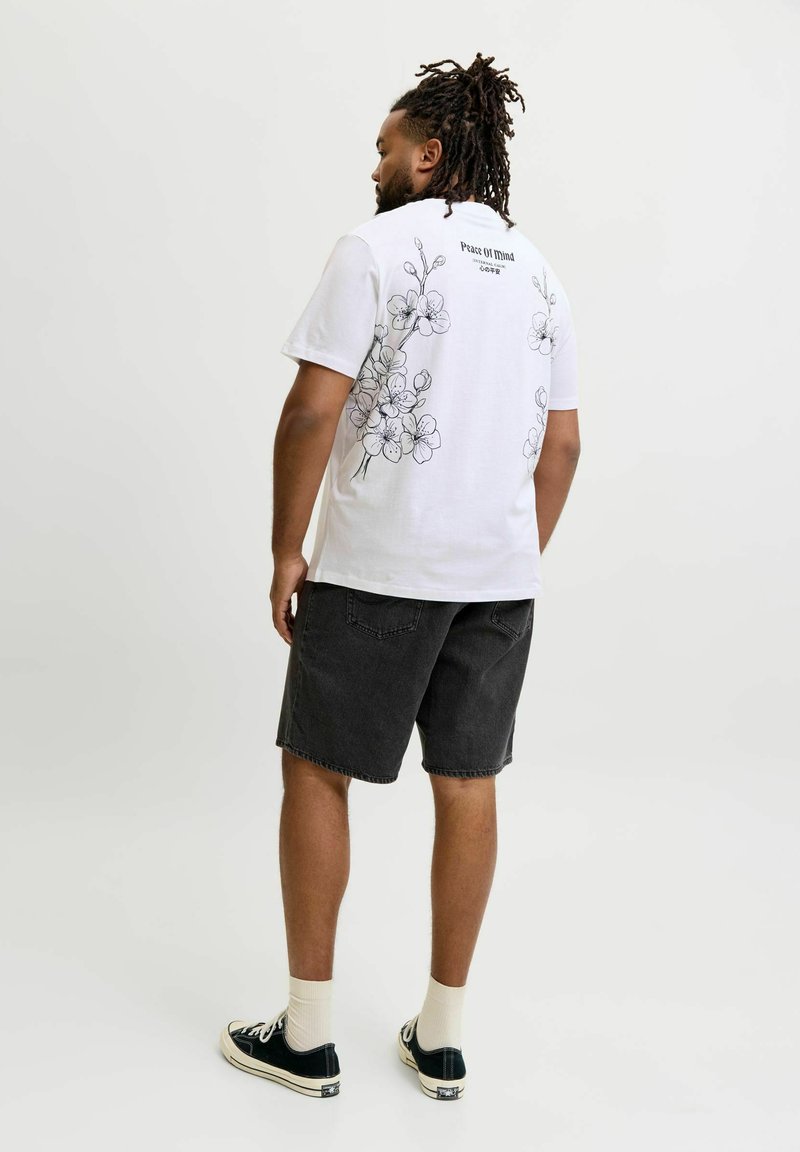 Άνδρας με dreadlocks που φοράει λευκό μπλουζάκι με floral σχέδιο, μαύρα denim σορτς, λευκές κάλτσες και μαύρα αθλητικά παπούτσια, στέκεται μπροστά σε απλό φόντο.