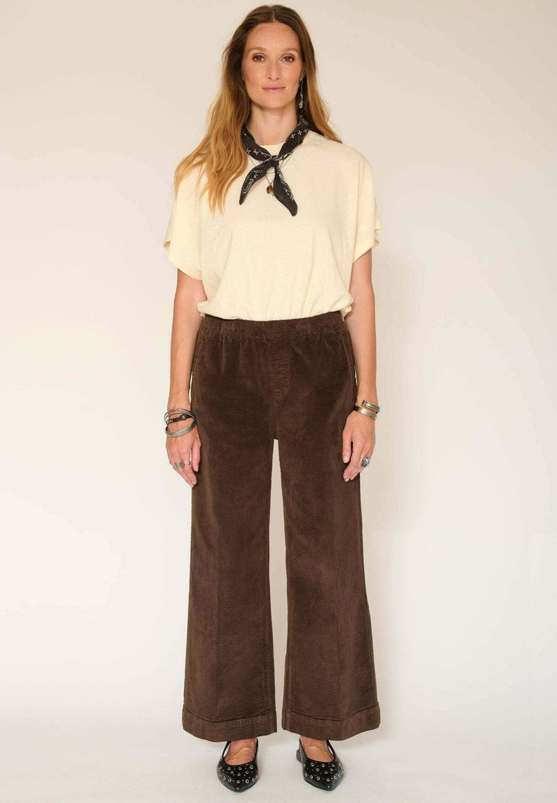 T-shirt oversized beige clair associé à un pantalon marron foncé en velours côtelé à jambes larges. Les accessoires incluent une écharpe noire et plusieurs bracelets en argent.