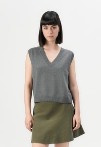 Woman with straight dark hair wearing a sleeveless gray V-neck knit top and an olive green A-line skirt against a plain background.