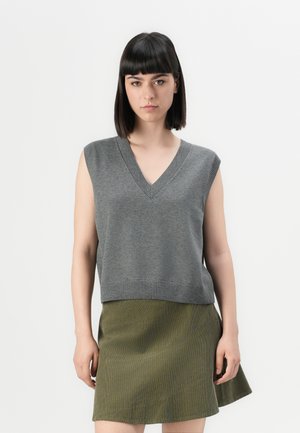 Vrouw met steil, donker haar die een mouwloze grijze V-hals gebreide top en een olijfgroene A-lijnrok draagt, tegen een effen achtergrond.