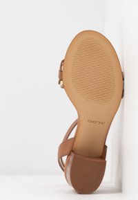 Sandale en cuir marron avec une semelle texturée, une bride arrière et un accent doré. La semelle présente des rainures horizontales pour une meilleure adhérence.