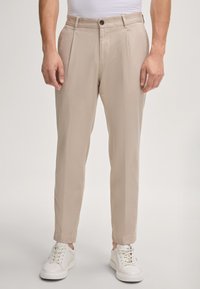 Pantaloni beige a gamba affusolata con pieghe frontali, realizzati in un tessuto leggero, abbinati a sneakers bianche. Dettaglio di chiusura con un singolo bottone.
