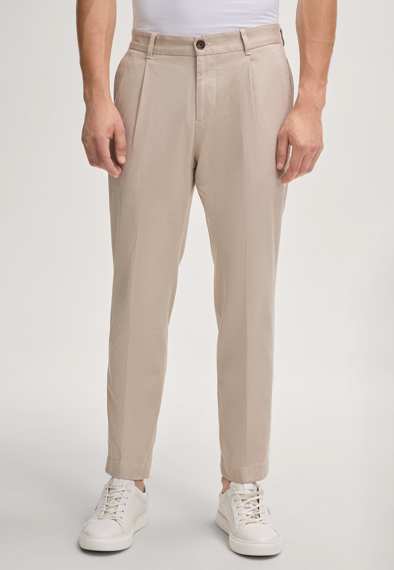Pantaloni beige a gamba affusolata con pieghe frontali, realizzati in un tessuto leggero, abbinati a sneakers bianche. Dettaglio di chiusura con un singolo bottone.