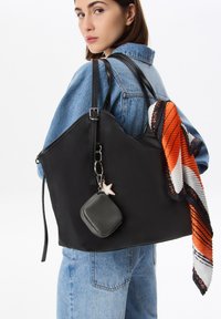 Femme portant une veste en denim, tenant un grand sac noir avec une petite pochette attachée et une écharpe orange-noir-blanche drapée sur la sangle du sac.