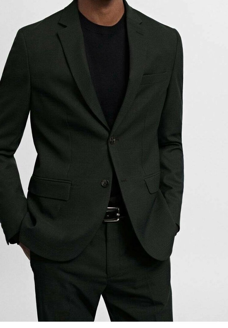 Homme portant un costume vert foncé avec une chemise noire et une ceinture noire, mains dans les poches, montré du cou jusqu'au milieu de la cuisse sur un fond uni.