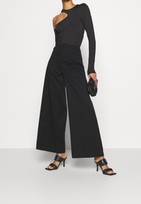 Czarny top z długim rękawem z wycięciami, w połączeniu z szerokimi czarnymi spodniami i czarnymi sandałami na obcasie. Dodatkiem jest mała czarna torebka.