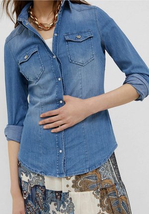 Femme portant une chemise en jean bleu clair aux manches retroussées et un collier en chaîne dorée, associée à une jupe à motif paisley.
