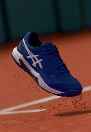 Blue athletic shoe with a mesh upper, white logo, textured surface, and rubber sole, set against a clay court backdrop.