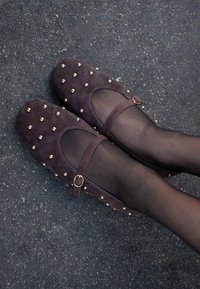 Ballerines en suédine violette foncée avec un bout rond, des clous dorés, et une sangle avec une boucle dorée, portées avec des collants noirs transparents sur une surface texturée.
