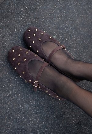 Ballerines en suédine violette foncée avec un bout rond, des clous dorés, et une sangle avec une boucle dorée, portées avec des collants noirs transparents sur une surface texturée.