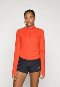 Svijetlo narančasta dugosveđa sportska majica s polu-zip ovratnikom, glatke teksture, i Nike logotipom. Uparen s crnim sportskim kratkim hlačama.