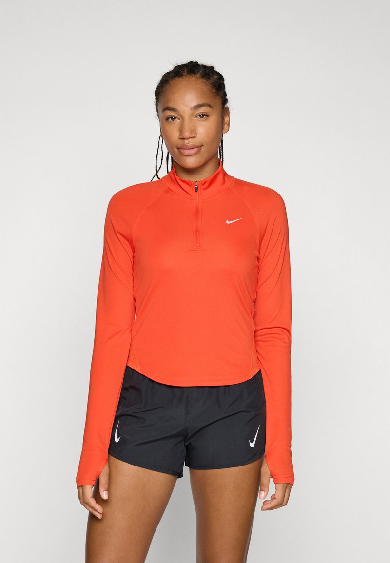 Svijetlo narančasta dugosveđa sportska majica s polu-zip ovratnikom, glatke teksture, i Nike logotipom. Uparen s crnim sportskim kratkim hlačama.