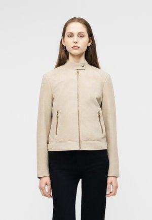 Žena s dlhými hnedými vlasmi, oblečená v béžovej mikine na zips s dvoma prednými vreckami na zips a čiernych nohaviciach, stojaca na jednolatom svetlom pozadí.