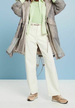 Lätt, beige kappa över en grön topp och randig skjorta. Vita vida byxor och beige sneakers fulländar outfiten.