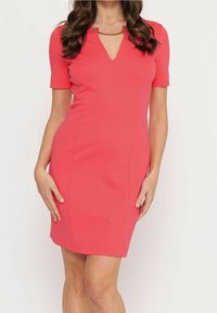 Žena v upnutých korálově růžových šatech ke kolenům s krátkými rukávy a zlatým řetízkem detailu u výstřihu.