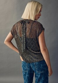 Gennemsigtig sort og gul top med korte ærmer, der har en sløjfe detaljer på ryggen; sat sammen med mønstrede blå jeans.