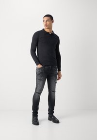 Sort langærmet polo shirt med krave, parret med slidte grå jeans med rifter. Sorte sko fuldender outfittet.