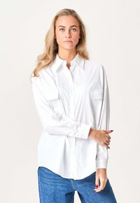 Blond kvinde med langt hår iført en oversized hvid skjorte og blå jeans, stående mod en ensartet lys baggrund.