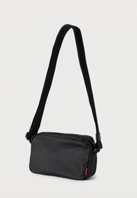 Melna taisnstūrveida plecu soma no fakturēta auduma. Tai ir regulējams siksniņas garums un rāvējslēdzēja aizdare ar sarkanu akcenta birku.