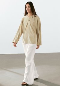 Beige Wildleder-Langarmshirt mit Knopfleiste, kombiniert mit weißen, weiten Hosen und schwarzen Flachsandalen, die über eine glatte Oberfläche laufen.