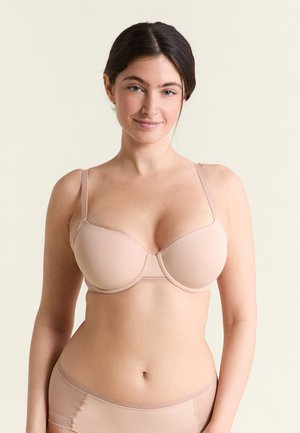 Mujer de cabello oscuro con sujetador acolchado beige y ropa interior a juego, de pie contra un fondo claro y liso, sonriendo suavemente.