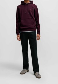 Burgunderröd hoodies med ficka fram och logotyp, parat med svarta byxor och grå sneakers. Enkel, avslappnad passform.