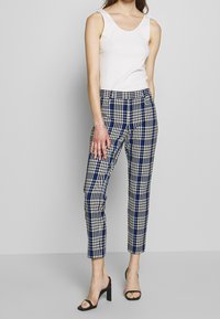 Pantaloni a scacchi con motivi blu e neri, vestibilità slim, lunghezza alla caviglia, abbinati a una canotta bianca e sandali neri con tacco.