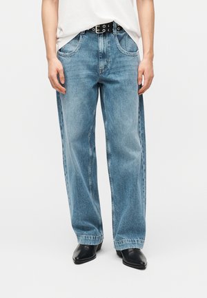 Osoba nosiaca svetlomodré rifle so širokými nohavicami, bielu zastrčenú košeľu, čierny opasok s očkami a čierne topánky s úzkou špičkou, stojaca pred bielym pozadím.