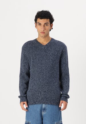 Navy blue knitted sweater with a V-neck, long sleeves, and textured fabric. Features ribbed cuffs and hem. Styled with light blue trousers.