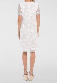 Femme portant une robe blanche en dentelle florale jusqu'aux genoux et des sandales compensées beiges, debout de dos sur un fond clair uni.