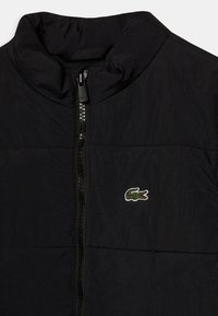 Svart quiltad jacka med hög krage, hel dragkedja och en liten grön lakert-logotyp på bröstet. Lättviktsmaterial med en strukturerad yta.