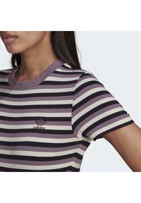 Kvinna som bär en lila, svart och vit randig Adidas t-shirt med en liten broderad logo på vänster bröst.