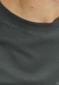 Tumši pelēks kokvilnas t-krekls ar apaļu kaklu un ribinātu apkakli. Gluda tekstūra ar minimālām šuvju detaļām. Tuvplāns skats, koncentrējoties uz kakla izgriezumu.