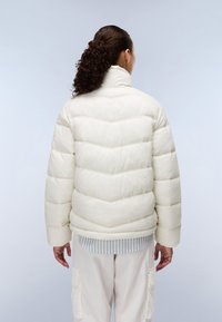Hvid dunjakke med chevron-quilting, høj krave og en let skinnende overflade; båret over en lyseblå og hvid stribet skjorte.
