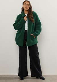 Zielona, puszysta kurtka zapinana na zamek z kołnierzem, z przednimi kieszeniami. Połączona z czarnymi spodniami o szerokich nogawkach i botkami za kostkę.