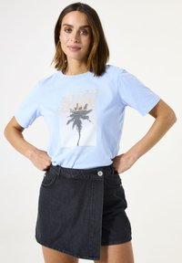 Lichtblauwe katoenen T-shirt met een palmbomenprint in zwart en goud. Gecombineerd met een zwarte denim rok met een asymmetrisch ontwerp.