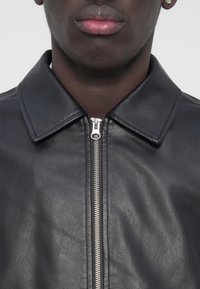 Veste en cuir noire avec un col à revers, dotée d'une fermeture éclair argentée et d'une texture lisse, mettant en valeur le design contemporain du vêtement.