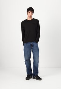 Chemise noire à manches longues avec un petit logo, associée à un jean bleu et des baskets noires. Design simple, coupe décontractée et couleurs unies.