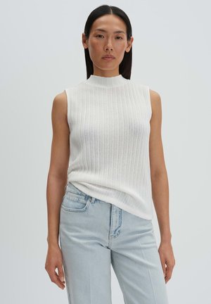 Vrouw met steil donker haar, gekleed in een mouwloos wit gebreid topje en lichtblauwe high-waisted jeans, staand tegen een effen achtergrond.