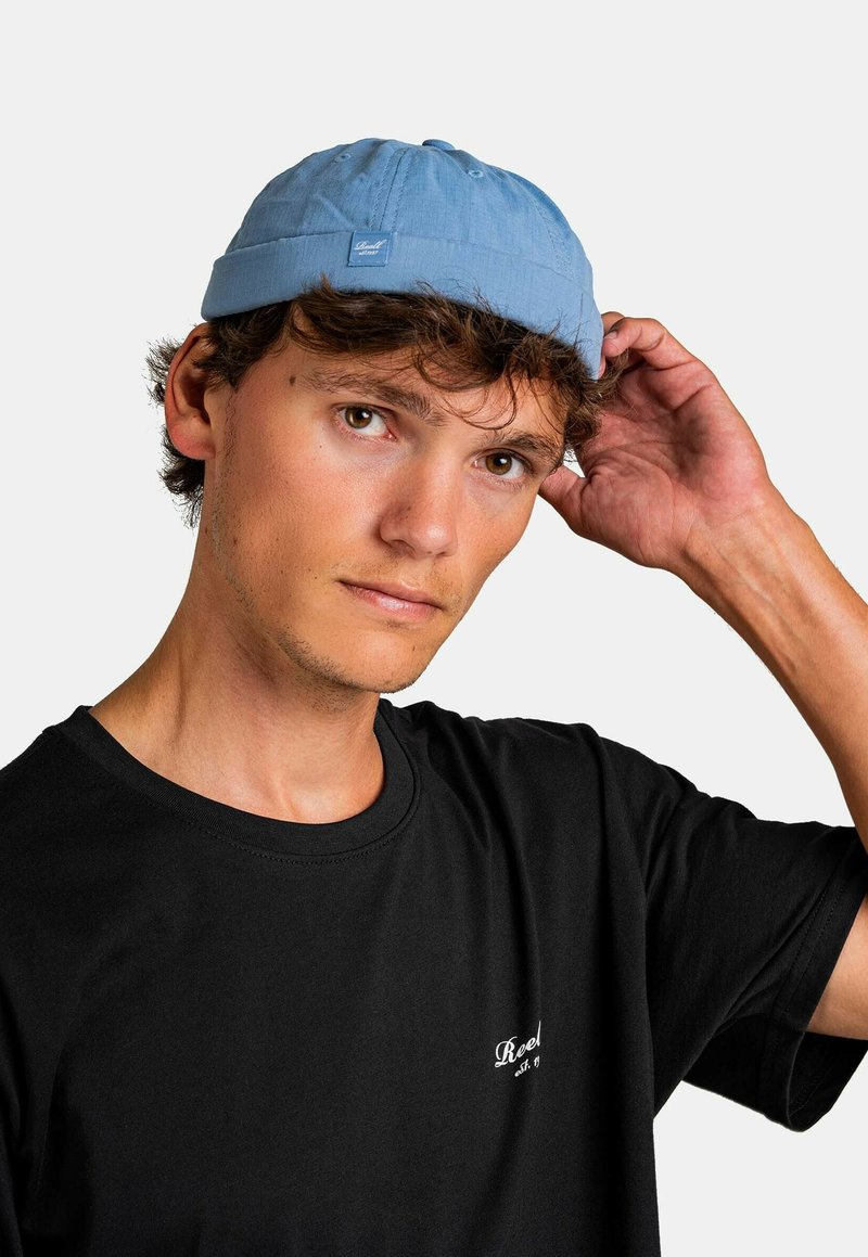 Gorra azul clara con una corona plana y una pequeña etiqueta de marca. Se lleva con una camiseta negra, que presenta un diseño sencillo y un ajuste casual.