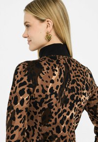 Blusa a stampa leopardo in marrone e nero con colletto alto; modello indossa orecchini a forma di cuore dorati; texture del tessuto liscia.