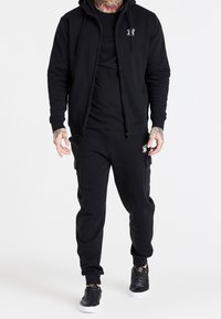 Černá kapucňová bunda na zip, černé tričko s kulatým výstřihem a černé tepláky, vše z hladké látky, doplněné černými teniskami. Bílé logo detaily.