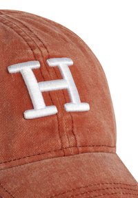 Cappello color ruggine realizzato in morbido tessuto, con una "H" ricamata in bianco ben visibile nella parte frontale. Presenta una superficie strutturata e cuciture visibili.