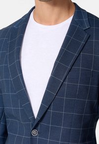 Blazer azul marino con un patrón de cuadros, tela texturizada, solapas con muesca, cierre de un solo botón y detalle de bolsillo, usado sobre una camisa blanca.