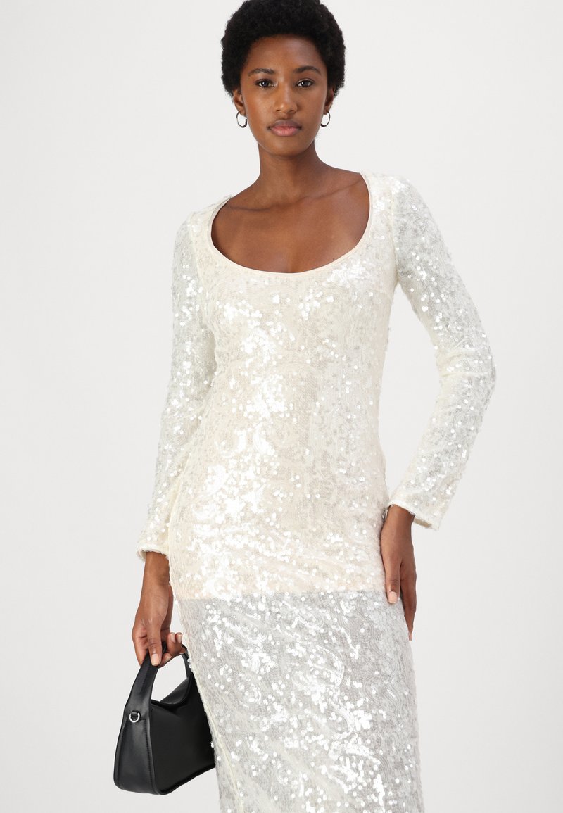 Długo rękawowa, cekinowa sukienka z okrągłym dekoltem w kolorze off-white i srebrnym. Teksturowana tkanina z dopasowaną sylwetką, w połączeniu z czarną torebką.