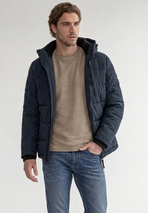 Homme portant une veste matelassée bleu marine sur une chemise beige et un jean bleu, debout, une main dans la poche, regardant sur le côté.