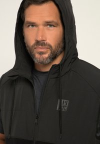 Schwarze Zip-Up-Weste mit Kapuze, die eine gesteppten Oberflächenstruktur aufweist, mit Kordeln und einem auffälligen Logo auf der Brust. Glattes Material mit einer sportlichen Passform.