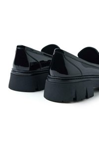 Zapatos de charol negro con suela de goma negra gruesa, que presentan un acabado suave y un ligero aumento en la parte posterior para mayor soporte.